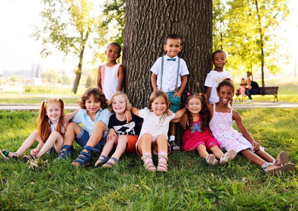 A group of small children sitting by a tree in the park. Unique Nannies